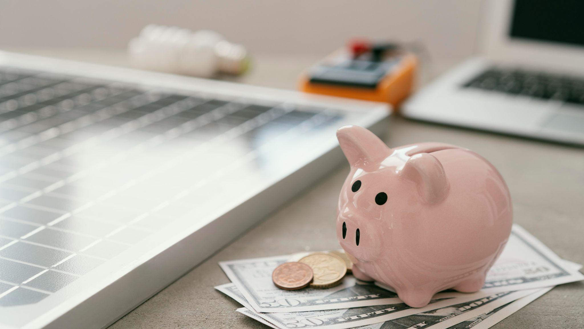 A pink piggy bank on banknotes beside a solar panel, calculator and laptop