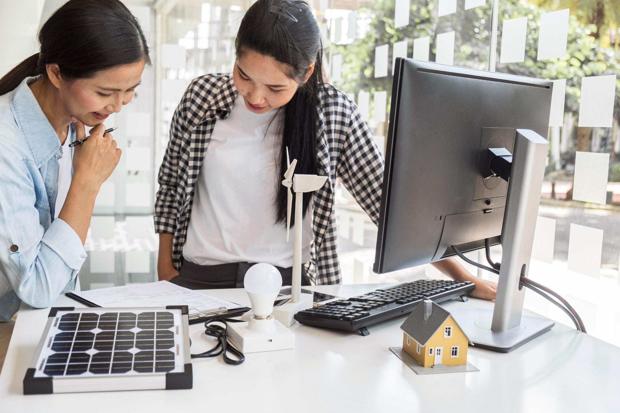 Two people reviewing solar and battery plans at a computer with a model house and wind turbine