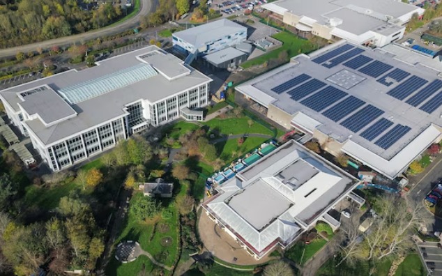 Aerial view of a large commercial building with a rooftop solar array beside office buildings and landscaped grounds