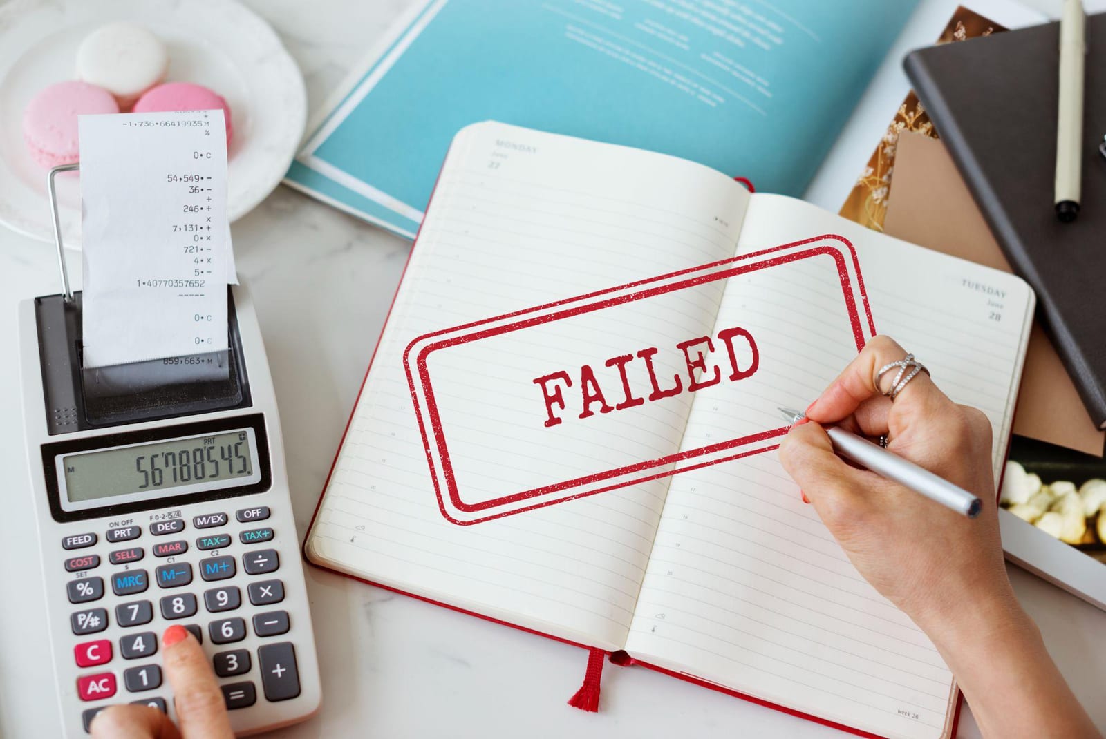 A hand writing in a diary stamped FAILED beside a printing calculator and receipt