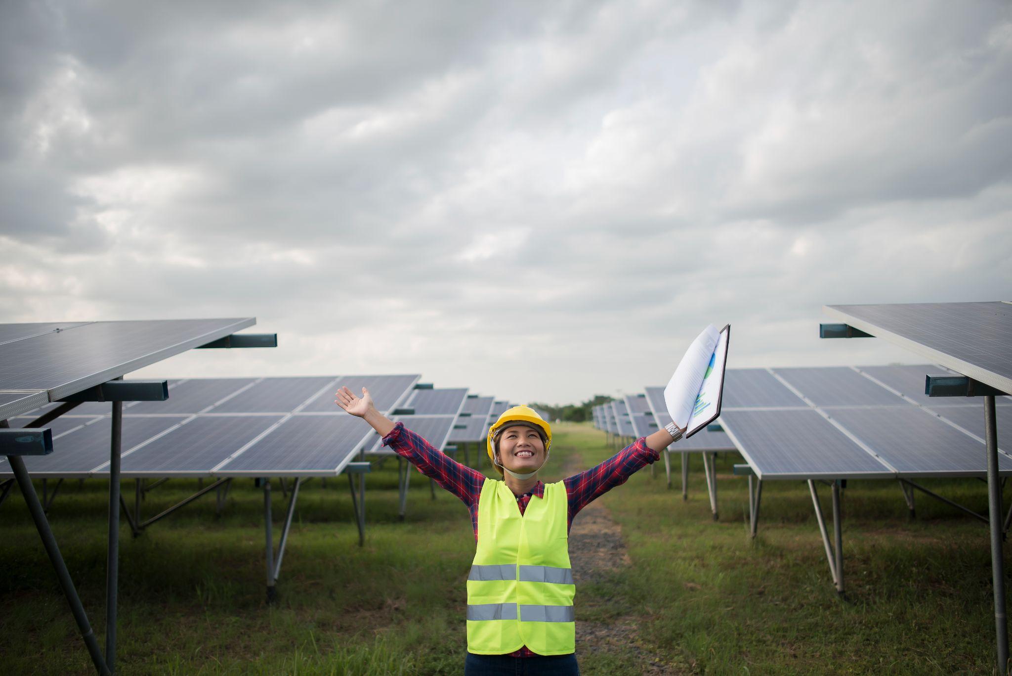 A solar engineer in a hi-vis vest standing with arms raised between rows of ground-mounted solar panels